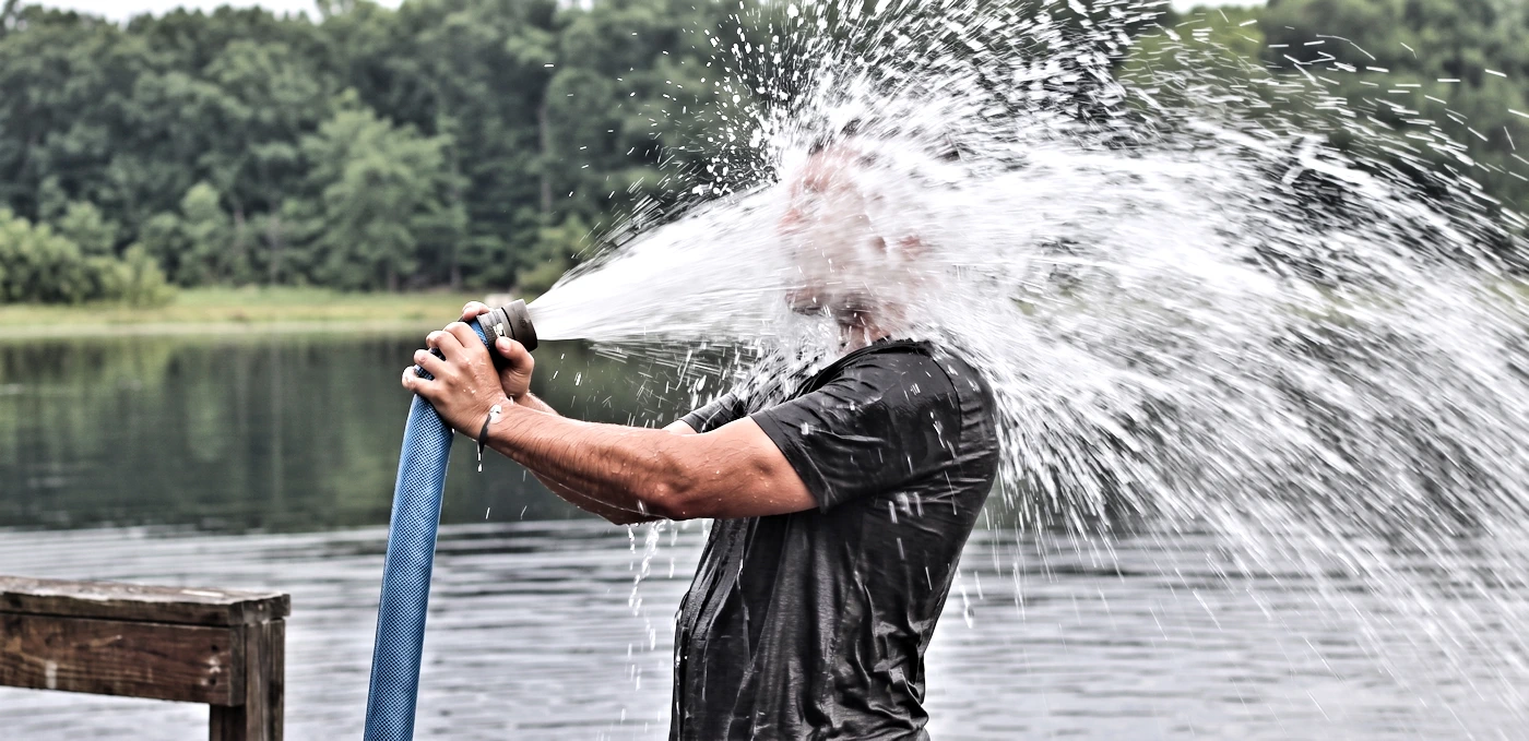 Side view profile of a man holding a high-pressure blue water hose blasting directly into his face next to a lake